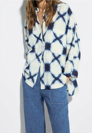 Femme portant une chemise blanche ample à boutons avec un motif tie-dye bleu marine et un pantalon en denim bleu taille haute à jambes larges.