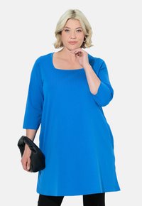 Blå, långärmad tunika med fyrkantig halsringning, gjord av mjukt tyg. Matchas med en svart texturerad clutch, bärs över svarta byxor.