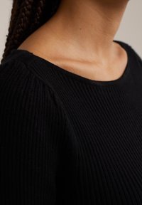 Pull en maille côtelée noire avec un large décolleté et un tissu texturé. Vue en gros plan mettant en valeur le matériau doux et extensible ainsi que les détails des épaules.