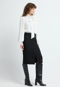 Blouse blanche avec un nœud papillon, manches bouffantes. Jupe noire avec une fente à l'avant. Bottes noires brillantes au genou avec un motif crocodile.