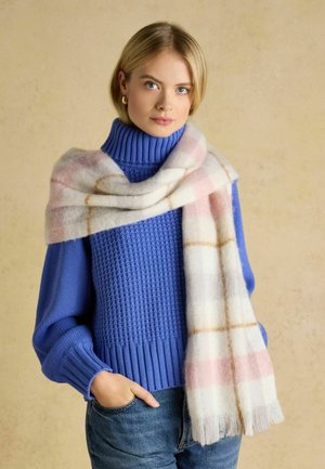 Blonde woman wearing a blue turtleneck sweater and a large white plaid scarf draped over her shoulders, with one hand in jeans pocket.