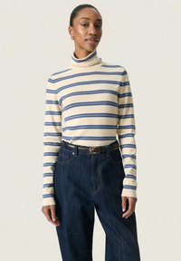 Mujer con un suéter de cuello alto color crema con rayas horizontales azules y jeans de tiro alto en azul oscuro con un cinturón negro.
