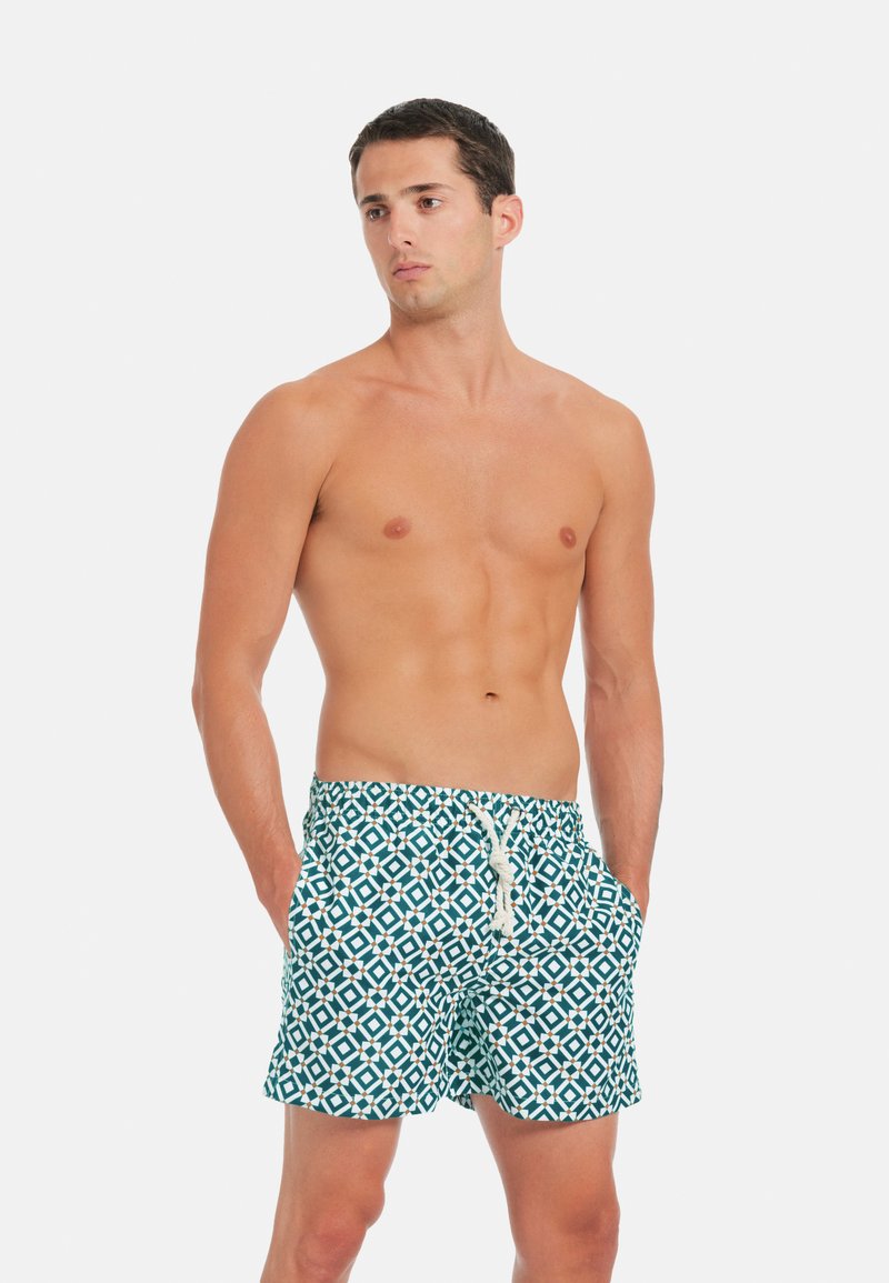 Jeune homme debout, les mains dans les poches, portant un short de bain à motifs géométriques bleu et blanc avec un cordon blanc.