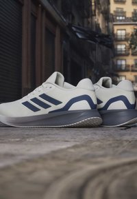 Weiße und marineblaue Laufschuhe mit drei marineblauen Streifen, auf einem Bürgersteig in der Stadt mit Gebäuden im Hintergrund platziert.