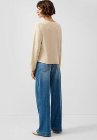 Frau mit kurzen braunen Haaren, die einen cremefarbenen Pullover und weit geschnittene blaue Jeans trägt, seitlich stehend vor einem schlichten hellgrauen Hintergrund.