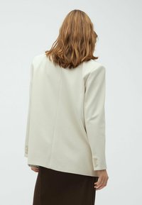 Persona con capelli castano chiaro di lunghezza media, che indossa un blazer oversize di colore avorio e una gonna scura, volta di spalle contro uno sfondo semplice.