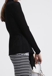 Černý top s dlouhým rukávem, přiléhavým pasem a peplum detailem, spárovaný se pruhovanou tužkovou sukní. Bílá kabelka držena v ruce.