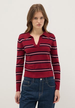 Jeune femme portant un pull à col rayé rouge, bleu marine et blanc à manches longues, associé à un jean denim bleu foncé, debout devant un fond uni.