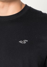 Černé tričko s krátkým rukávem vyrobené z bavlny s kulatým výstřihem a malým bílým logem ptáka na levé hrudi. Měkká textura, ležérní design.