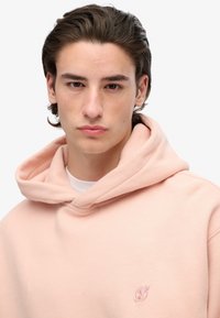 Sudadera con capucha rosa hecha de una tela suave, con un pequeño logo bordado en el lado izquierdo del pecho, corte relajado y capucha ajustable con cordón.