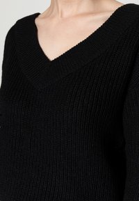 Close-up van een persoon die een zwarte geribbelde gebreide trui met een V-hals draagt, waarbij een deel van de nek en de bovenborst zichtbaar is.