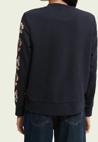 Granatowy sweter z długim rękawem, z haftem kwiatowym na lewym rękawie oraz żebrowanym wykończeniem u dołu i na rękawach. Gładki, średniej grubości materiał.