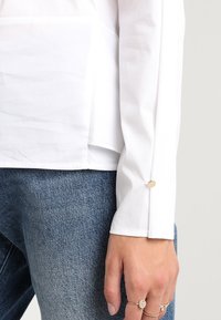 Camicia bianca a maniche lunghe con taglio dritto, caratterizzata da dettagli con bottoni sui polsini, abbinata a jeans in denim blu chiaro.