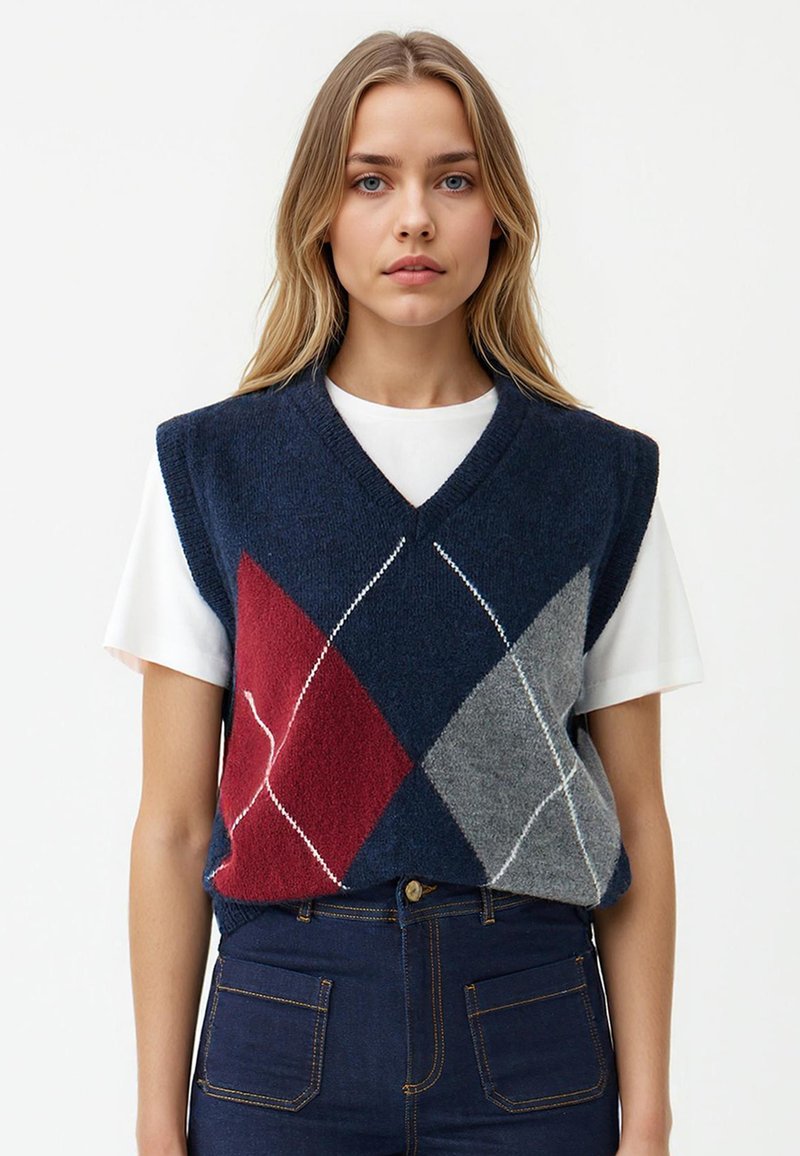 Maglione senza maniche a V blu navy con motivi a rombi rossi, grigi e bianchi. Indossato sopra una t-shirt bianca, abbinato a jeans in denim scuro.