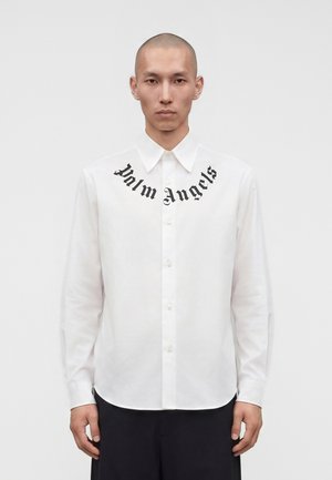 Bărbat cu capul ras, purtând o cămașă albă cu nasturi, cu inscripția "Palm Angels" în font gotic negru pe piept și pantaloni negri.