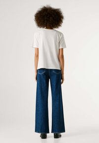 Maglietta bianca di cotone con maniche corte abbinata a jeans di denim blu scuro a gamba larga. Calzature nere visibili nella parte inferiore. Design semplice, senza motivi.