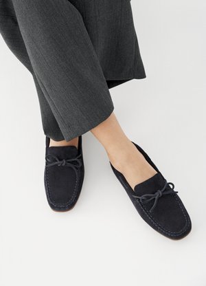 Άτομο που φοράει σκούρα γκρι παντελόνια και μπλε ναυτικό μοκασίνια από σουέντ με λεπτομέρεια φιόγκου, στέκεται σε λευκή επιφάνεια.