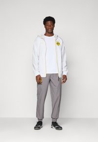 Homme portant une veste blanche Puma avec le logo jaune de Borussia Dortmund, une chemise blanche, un pantalon gris et des chaussures de sport noires, se tenant devant un fond uni.