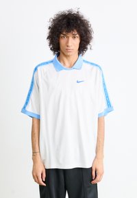 Polo de manga curta da Nike em branco, com gola e punhos em azul claro, apresentando riscas laterais azuis e um pequeno logótipo no peito.
