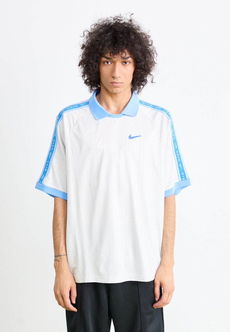 Polo de manga curta da Nike em branco, com gola e punhos em azul claro, apresentando riscas laterais azuis e um pequeno logótipo no peito.