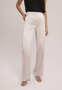 Spodnie o wysokim stanie, zwężające się ku dołowi z szerokimi nogawkami, wykonane z miękkiego, błyszczącego kremowego materiału. Posiadają kieszenie z przodu oraz gładką talię, w zestawie z przezroczystymi obcasami.