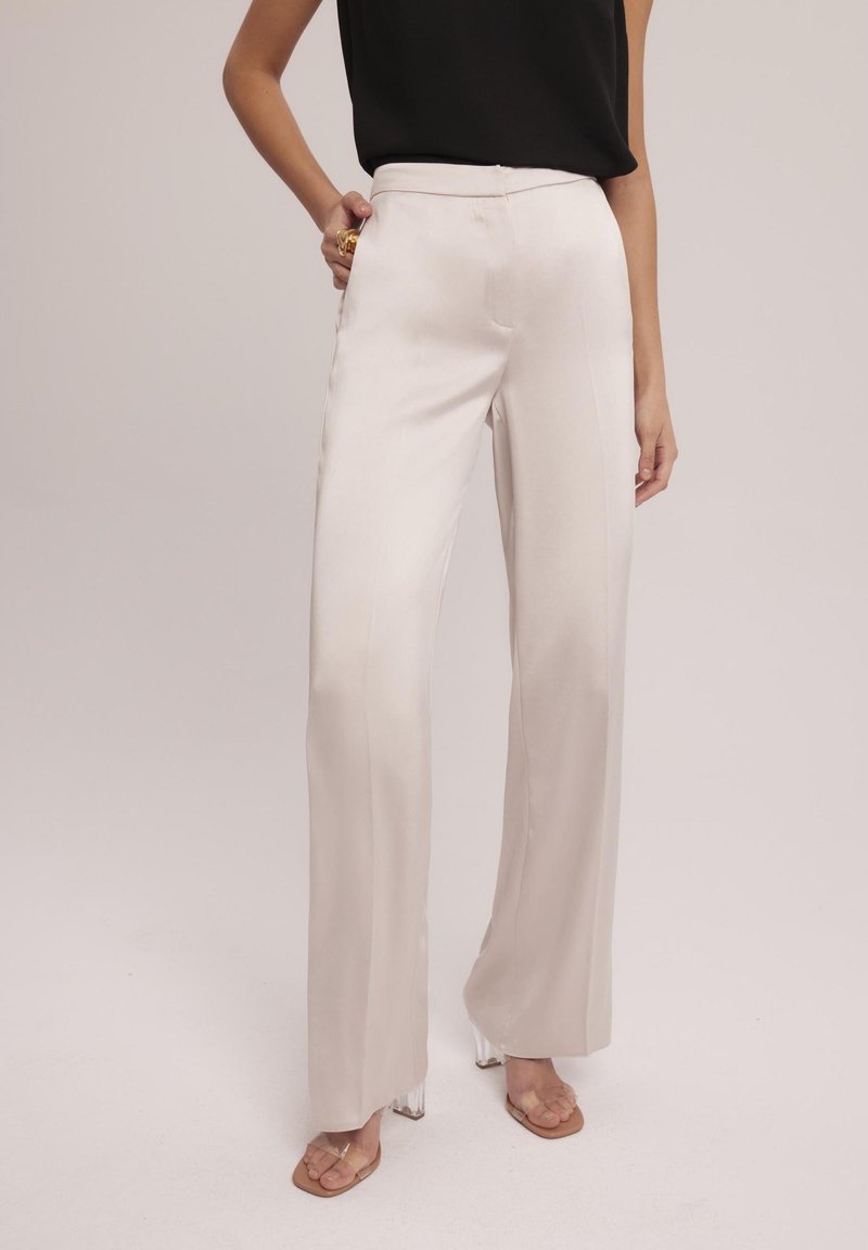 Spodnie o wysokim stanie, zwężające się ku dołowi z szerokimi nogawkami, wykonane z miękkiego, błyszczącego kremowego materiału. Posiadają kieszenie z przodu oraz gładką talię, w zestawie z przezroczystymi obcasami.