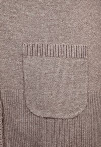 Jasnobrązowy sweter z dzianiny, z kwadratową kieszenią o prążkowanych krawędziach i gładką fakturą. Tkanina ma delikatny połysk.