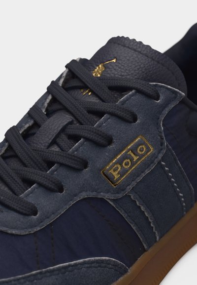Σκούρο μπλε αθλητικό παπούτσι με σουέντ λεπτομέρειες, σκούρα κορδόνια, χρυσό έμβλημα με το λογότυπο «Polo» και ραμμένες λεπτομέρειες σε καφέ σόλα.