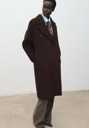 Cappotto doppiopetto in lana bordeaux con grandi bottoni neri, abbinato a pantaloni a quadri grigi e una camicia azzurra chiara. Texture liscia.