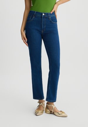 Jeans in denim blu con vita alta, taglio dritto e tasche posteriori, abbinati a scarpe piatte dorate con cinturini incrociati e dettagli.