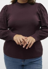 Donna che indossa un maglione viola scuro con maniche a sbuffo testurizzate e jeans blu, mani incrociate davanti all'altezza della vita.