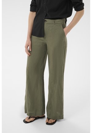 Persona vistiendo pantalones de lino verde oliva de pierna ancha, camisa negra abotonada con las mangas remangadas y chanclas negras, de pie con las manos en los bolsillos.