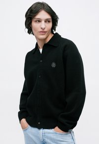 Cardigan negru tricotat, cu un guler la gât, nasturi pe față, manșete strâmte și un logo brodat pe partea stângă.