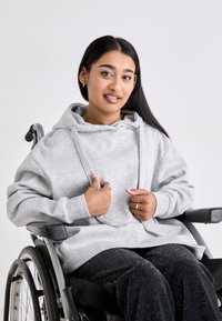 Grå, overdimensjonert hettegenser med kengurulomme og snor i hetten, iført over svarte, glitrende joggebukser. Modell sitter i rullestol.