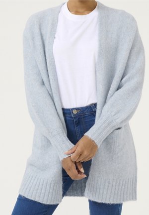 Person in einer hellblauen, flauschigen Strickjacke über einem weißen T-Shirt und blauen Jeans, die Hände vor dem Körper verschränkt, neutraler Hintergrund.