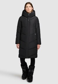 Frau trägt einen langen, schwarzen, gesteppte Wintermantel mit hohem Kragen, Reißverschluss und Druckknopfverschluss, schwarze Leggings und schwarze Winterstiefel.