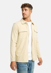 Ung mand iført en beige kordfløjljakke over en hvid skjorte og blå jeans, der ser til sin venstre side mod en ensfarvet baggrund.