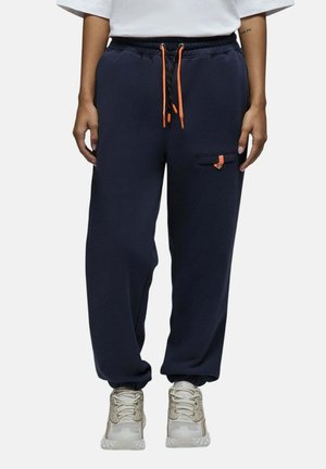Personne portant un pantalon de jogging bleu marine avec des cordons orange et un t-shirt blanc, debout devant un fond uni.