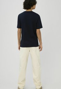 Personne aux cheveux bouclés portant un t-shirt noir à manches courtes et un pantalon couleur crème, debout devant un fond clair uni.