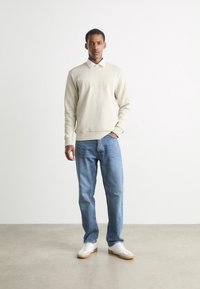 Sudadera beige con logo "BOSS" en relieve, cuello blanco, jeans azul claro y zapatillas blancas con suelas marrones, de pie sobre un fondo neutro.