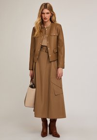 Brązowa skórzana kurtka z zatrzaskami, zestawiona z pasującą długą spódnicą z kieszeniami. Modelka trzyma jasnobeżową torebkę i nosi brązowe botki.