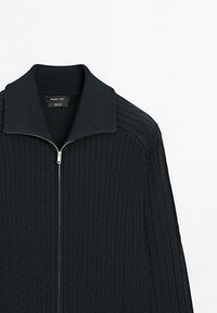 Maglione nero a maglia intrecciata con zip, colletto alto, maniche lunghe e una cerniera argentata visibile.