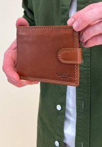 Hnědá kožená peněženka s jemnou texturou, vybavená zapínáním na knoflík a bílým šitím. Na přední straně je vyražen název značky.