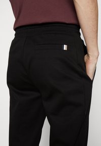Black sweatpants featuring a back pocket with a horizontal stripe in blue, white, and beige. Smooth fabric and elastic waistband design.