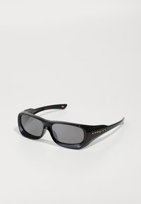 Óculos de sol retangulares pretos da Oakley com lentes escuras, logótipo dourado ao lado e braçadeiras ligeiramente curvadas, pousados sobre uma superfície branca.
