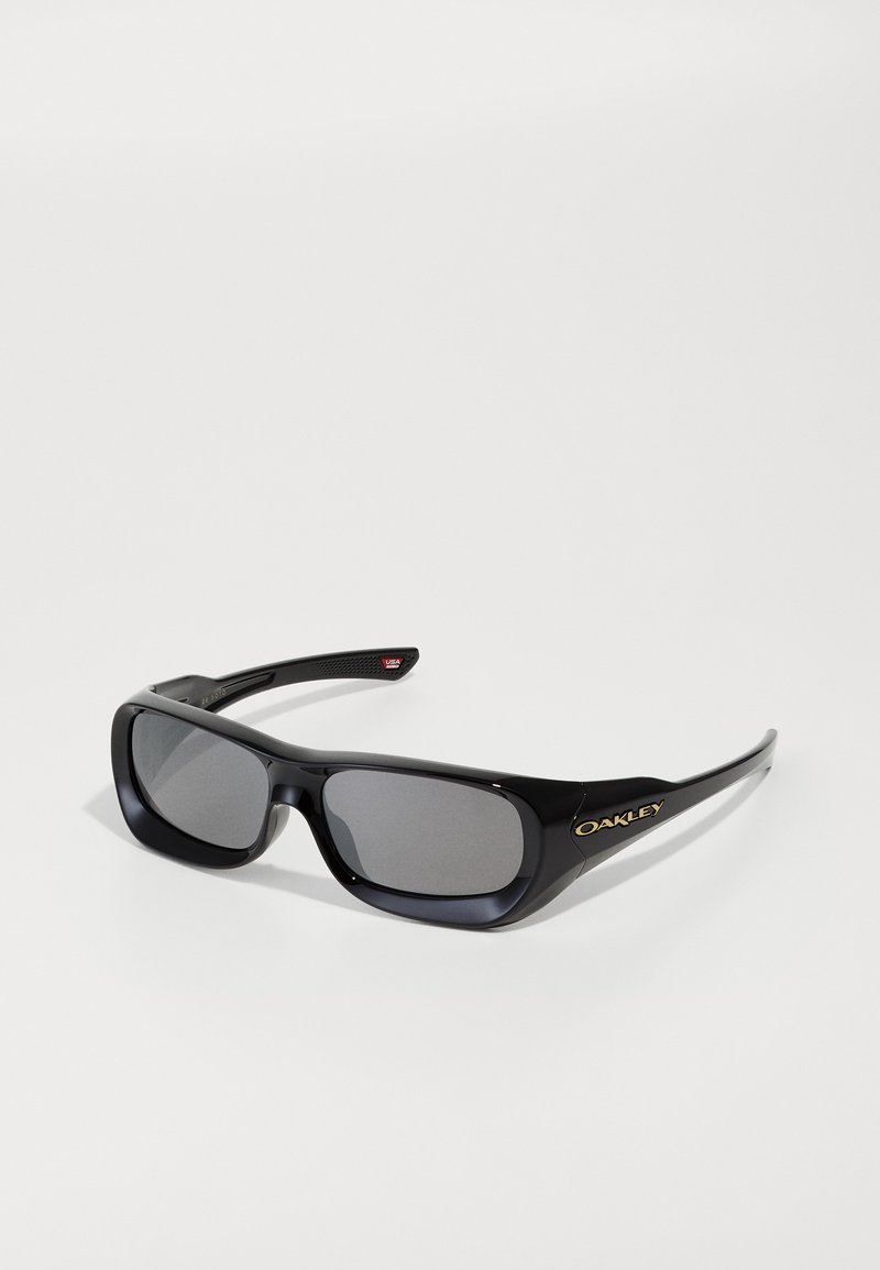 Μαύρα ορθογώνια γυαλιά ηλίου Oakley με σκούρους φακούς, χρυσό λογότυπο στο πλάι και ελαφρώς καμπυλωτά βραχίονες που είναι τοποθετημένα σε μια λευκή επιφάνεια.