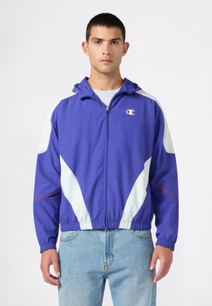 Man staat met het gezicht naar voren, draagt een blauw-witte Champion-jas met rits en capuchon en lichtblauwe spijkerbroek, op een witte achtergrond.