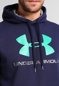 Felpa blu navy realizzata in tessuto morbido, con un grande logo Under Armour turchese e testo grigio, dotata di cordini e tasca centrale.