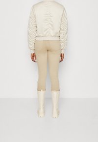 Osoba ubrana w beżowe obcisłe spodnie, kremową bomberkę i kremowe kozaki do kolan, stojąca na tle jednolitej białej ściany.