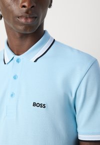 Mężczyzna ubrany w jasnoniebieską koszulę polo z białymi i czarnymi akcentami, z haftowanym logo "BOSS" po lewej stronie klatki piersiowej, patrzy na wprost z neutralnym wyrazem ust.